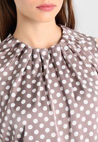 Haut sans manches marron avec des pois blancs et un col plissé. Confectionné dans un tissu lisse et léger.