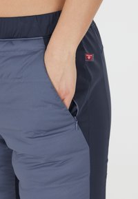 Marina och grå byxor med en ficka med dragkedja, elastisk midja och en Primaloft-logotyp. Texturerat material erbjuder en kombination av komfort och funktion.