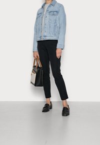 Lichtblauwe denimjack met twee borstzakken, grijs shirt eronder, zwarte slim-fit broek en zwarte instappers. Houdt een zwarte handtas vast.