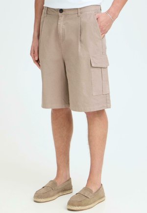 Beige cargoshort tot op de knie met zijflapzak, gedragen met beige instapschoenen en een wit shirt, persoon staand met één hand in de zak.