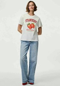 T-shirt branca de algodão com um gráfico vermelho "FESTIVAL DO MORANGO" e duas ilustrações de morangos; combinado com jeans azul claro.