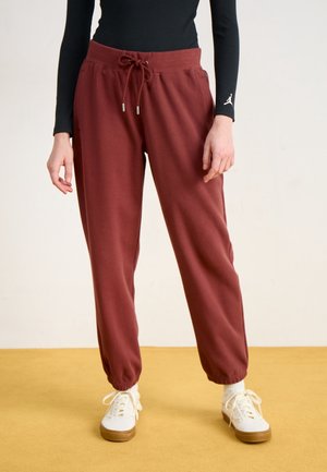 Personne portant un pantalon de jogging bordeaux texturé avec des poignets élastiques et des baskets blanches, debout sur un sol jaune.