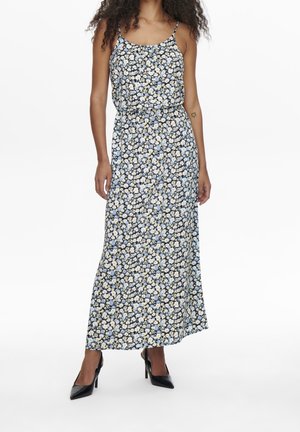 Frau trägt ein langes, ärmelloses Blumenkleid mit blauen, weißen und gelben Blumen, kombiniert mit schwarzen spitzen High Heels.