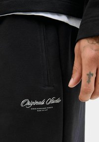 Pantaloni de trening negri cu o textură netedă. Prezintă un logo brodat "Originals Studio" și detalii cu buzunare. Mână cu un tatuaj vizibil.