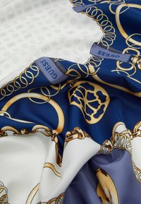 Foulard in seta con un design blu navy e bianco, arricchito da motivi a catena dorati e un'etichetta "GUESS" in blu. Texture liscia e si drappeggia elegantemente.