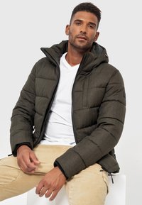 Dunkelgrüne gepolsterte Jacke mit Reißverschluss, Kapuze und Seitentaschen. Strukturierter Stoff und elastische Bündchen. Über einem weißen T-Shirt getragen.