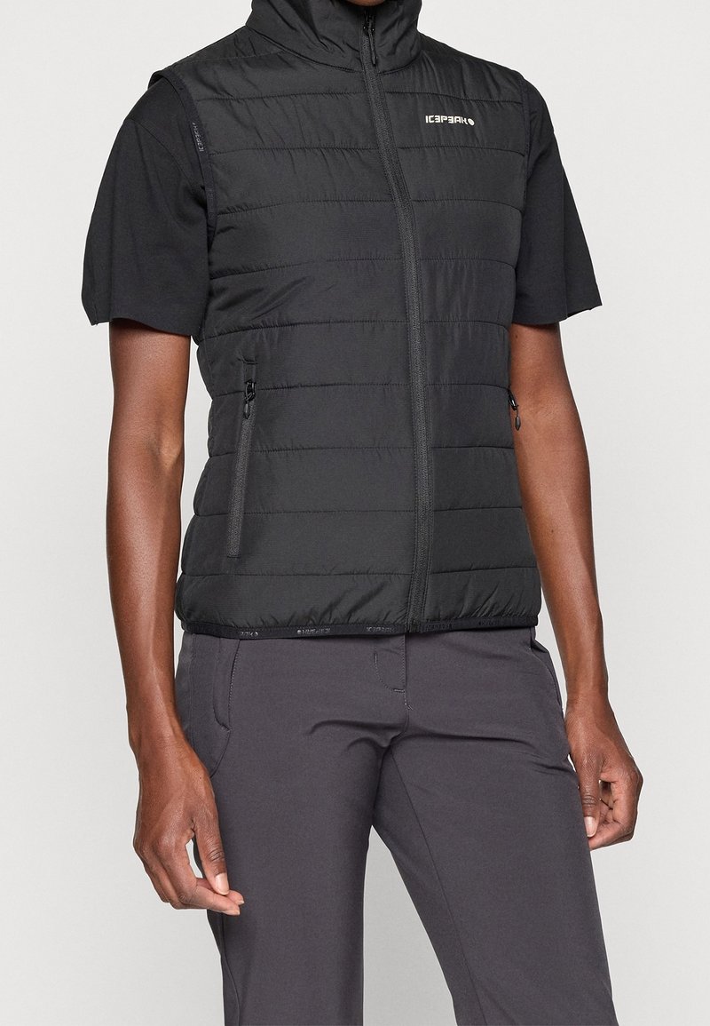 Personne portant une veste matelassée noire sans manches avec une fermeture éclair et deux poches latérales zippées, par-dessus un t-shirt noir à manches courtes et un pantalon foncé.