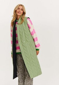 Grünes, gestepptes langes Weste mit schwarzem Innenfutter, ausgestattet mit Druckknöpfen und Seitentaschen, über einem mehrfarbigen gestreiften Pullover und Hosen im Leopardenmuster getragen.