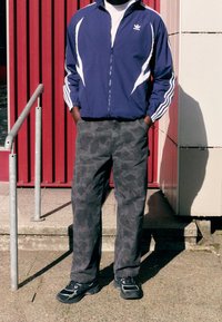 Blue zip-up jacket with white stripes, gray camo pants, and black sneakers. Standing on steps with a red wall in the background.
