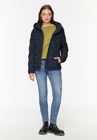 Mornarska puffer jakna s kapuco, zadrgo spredaj in stranskimi žepi, oblečena nad zeleno majico, v kombinaciji z svetlo modrimi strganimi denim hlačami in črnimi škornji.