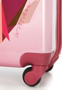 Valise rigide rose avec des motifs géométriques dans des nuances de rose et de brun, dotée d'une fermeture éclair rouge et de roues noires pour la mobilité.