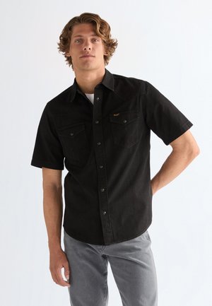 Noor mees laineliste juustega, kes kannab musta lühikeste varrukatega nööbitavat särki ja halli pükse, seistes ühe käega puusal.