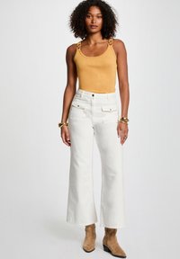 Yellow, sleeveless top with decorative hardware on straps, paired with white wide-leg pants. Model wears brown ankle boots.