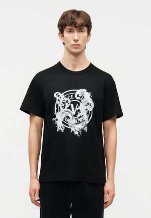 Jonge man die een zwart T-shirt draagt met een wit, sierlijk cirkelvormig "Versace Jeans Couture" logo in het midden van de voorkant, staande tegen een effen achtergrond.