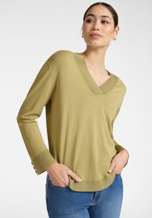 Mujer de cabello oscuro con un suéter de cuello en V de color verde oliva claro con puños y dobladillo acanalados, lleva jeans azules, manos en los bolsillos, mirando de lado.