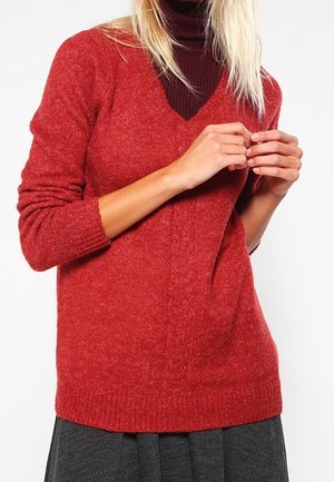 Femme portant un pull rouge en maille à col en V par-dessus un col roulé bordeaux foncé, les mains légèrement jointes et une jupe grise visible en dessous.