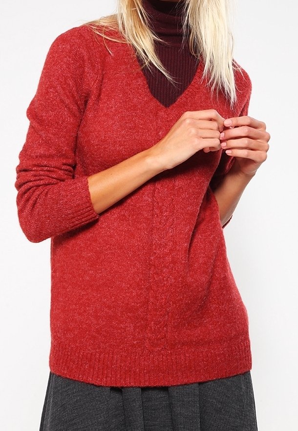 Femme portant un pull rouge en maille à col en V par-dessus un col roulé bordeaux foncé, les mains légèrement jointes et une jupe grise visible en dessous.