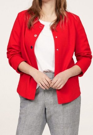 Femme portant un cardigan rouge vif boutonné sur une chemise blanche et un pantalon gris, les mains jointes à la taille.