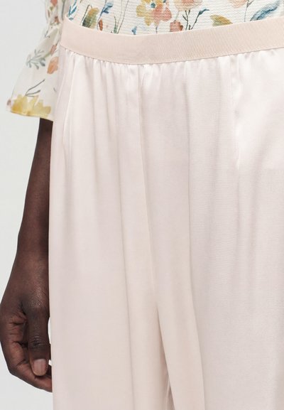 Κοντινό πλάνο ενός ατόμου που φοράει ανοιχτό ροζ σατέν παντελόνια με ελαστική μέση και μια φλοράλ μπλούζα με παστέλ χρώματα.
