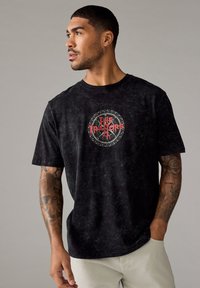 Zwarte katoenen t-shirt met een circulaire grafiek met "The Traitors" in het rood, textuurstof met een vervaagd uiterlijk, ronde hals, korte mouwen.