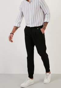 Černé zúžené kalhoty s záhyby, v kombinaci s bílou košilí s proužky a bílými teniskami. Košile má vyhrnuté rukávy.