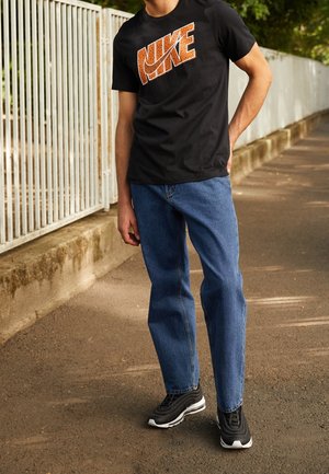 T-shirt noir avec logo "NIKE" orange, jean denim bleu, et baskets noires avec semelle blanche. Personne debout sur une surface pavée.