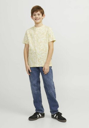 Geel T-shirt met korte mouwen en wit bladdessin, gecombineerd met blauwe denim jeans en zwarte sneakers met witte accenten.