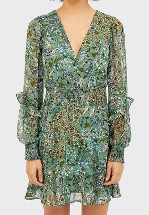 Lange jurk met groene bloemenprint, lange mouwen, V-hals, geplooide taille en gerimpelde manchetten, gedragen door een persoon waarvan het hoofd en de bovenbenen net boven de dijen niet zichtbaar zijn.