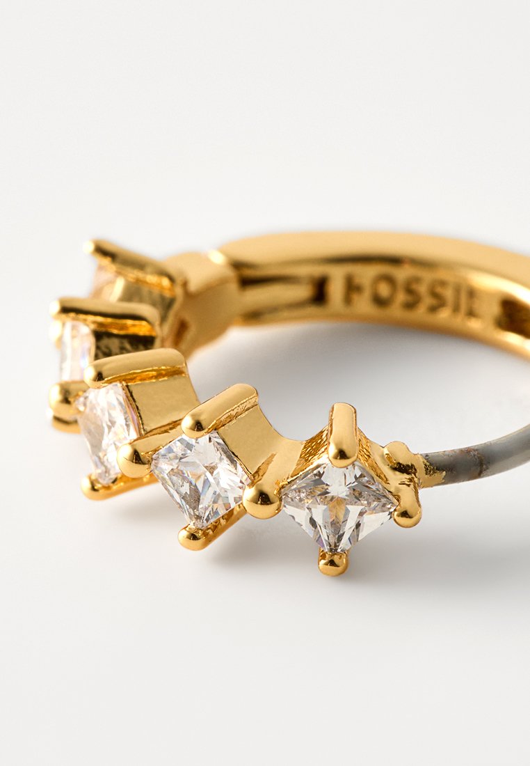 Bague en or ornée de quatre pierres carrées transparentes serties sur griffes. L'anneau est lisse avec une finition polie, affichant une gravure de marque.