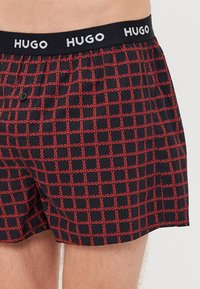 Boxers noirs avec un motif répétitif du logo rouge 'HUGO', taille élastique avec le texte 'HUGO' et une fermeture à bouton à l'avant.