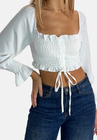 Witte cropped top met geplooide structuur, vierkante halslijn en koord aan de voorkant. Heeft wijde mouwen en een gerimpelde zoom. Gecombineerd met donkere jeans.