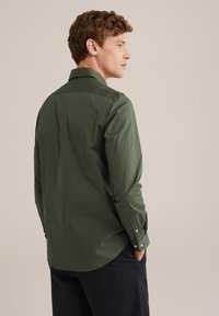 Hombre vistiendo una camisa de manga larga verde oliva y pantalones negros, de pie con las manos en los bolsillos, mirando de lado contra un fondo beige.