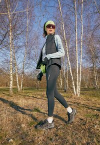 Gilet nero senza maniche sopra una maglietta a maniche lunghe azzurro chiaro, abbinato a leggins neri e scarpe da ginnastica scure. Cerchietto verde e guanti.