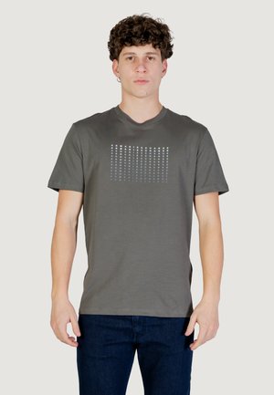 Jeune homme aux cheveux bouclés portant un t-shirt gris Armani Exchange avec un logo répété et un jean bleu foncé, debout devant un fond uni.