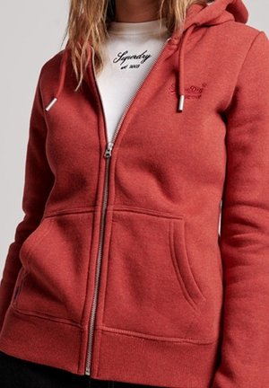Žena nosící červenou mikinu na zip přes bílé tričko značky Superdry, s rukama opřenýma poblíž předních kapes.