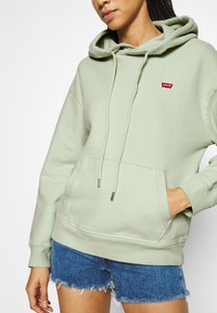 Světle zelená mikina z měkkého materiálu, s přední kapsou, šňůrkami s kovovými špičkami a malým červeným logem na hrudi. V kombinaci s modrými džínovými šortkami.