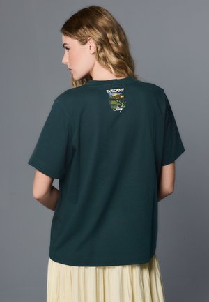 Donna con capelli mossi vista di spalle, indossa una maglietta verde scuro con una piccola grafica del paesaggio della Toscana, Italia, e testo vicino alla scollatura.