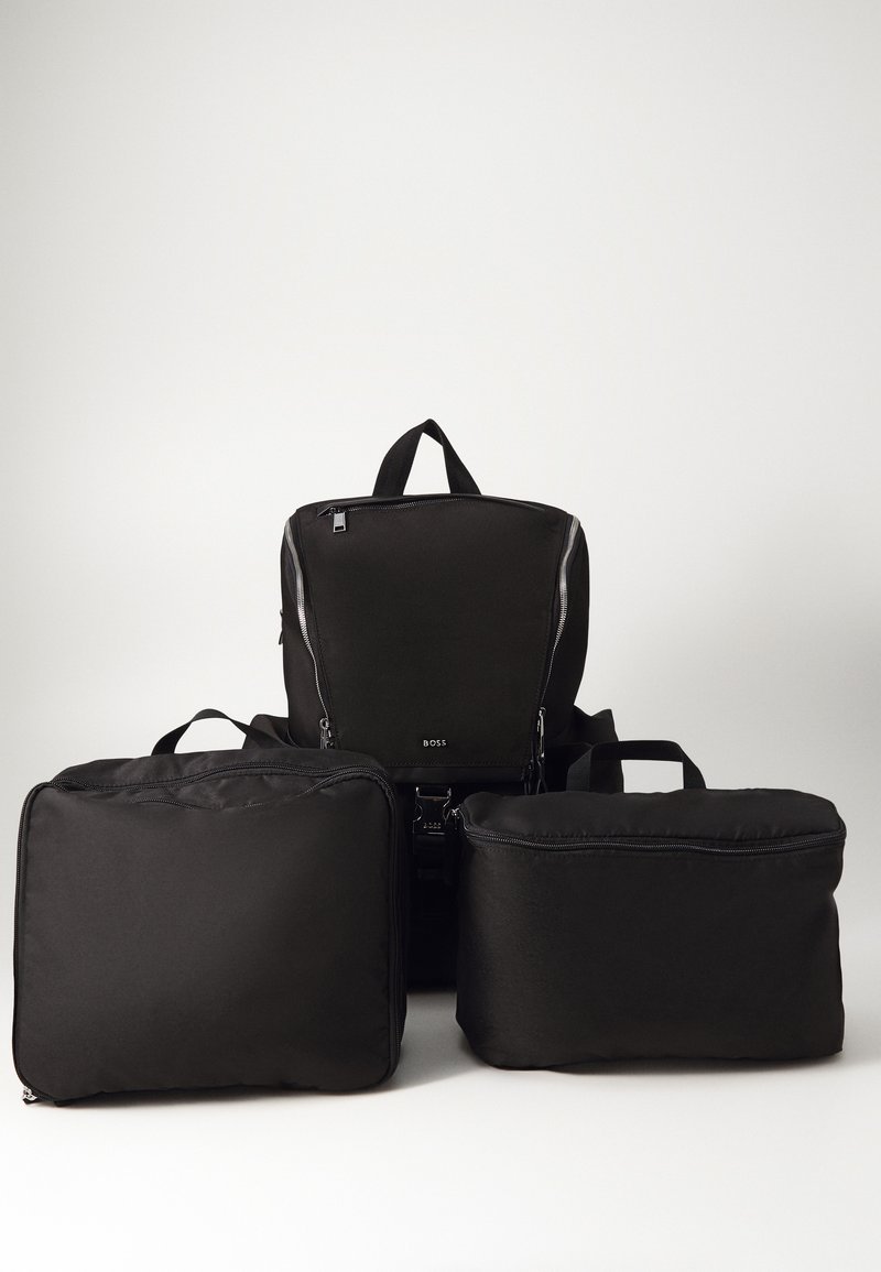 Un sac à dos noir et deux pochettes noires en tissu, présentant des textures lisses, des fermetures éclair et des poignées, disposés en formation empilée.