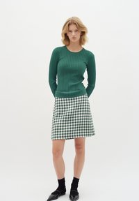 Zöld csíkozott, hosszú ujjú felső, amelyet zöld és fehér houndstooth mintás szoknyával párosítanak. Fekete bokazokni és hegyes orrú lapos cipő egészíti ki az öltözéket.
