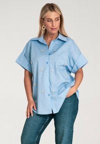Jasnoniebieska koszula z krótkim rękawem z guzikiem, ozdobiona haftem kwiatowym i dekoracyjnymi detalami. Wykonana z miękkiej bawełny, luźny krój.