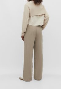 Beżowy krótki sweter z długimi rękawami nałożony na jasną bluzkę, w komplecie z spodniami o wysokim stanie i szerokich nogawkach w delikatnym, neutralnym odcieniu.