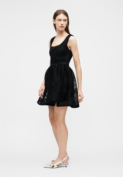 Γυναίκα με αμάνικο μαύρο δαντελένιο φόρεμα με φουσκωτή φούστα, φορώντας λευκές χαμηλές γόβες με λουράκι στην πλάτη, γυρισμένη αριστερά πάνω σε απλό φόντο.