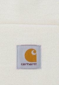 White knit beanie with a ribbed texture. Includes a gray patch featuring a yellow logo and the brand name "carhartt" in brown.
