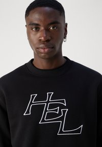 Mustvalg musta suletoodetiga, millel on ees valgete tikanditega tekst "HEL". Kanga pind näib siledana, tavaline lõige ja ribaga krae.