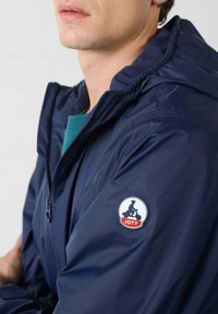 Waterdichte jas in marineblauw met capuchon, voorzien van een logopatch op de linkerarm. Het materiaal oogt glad en licht van gewicht.