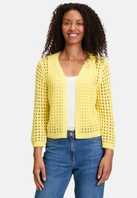 Cardigan jaune en tricot avec un design ajouré, poches avant et texture légère, associé à un haut blanc et un jean en denim bleu.