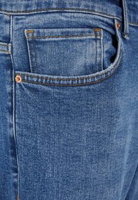 Jean en denim bleu clair avec une poche arrière, des coutures dorées et une rivet en métal. Le tissu a une surface légèrement texturée.
