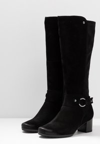 Botas de ante negras hasta la rodilla con un tacón robusto, que cuentan con un acento circular y una cremallera lateral para facilitar su uso. Textura suave y diseño ajustado.