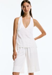 Top halter in lino bianco con scollatura a V profonda e fit morbido, abbinato a pantaloni corti bianchi fino al ginocchio. Design semplice, aspetto minimalista.