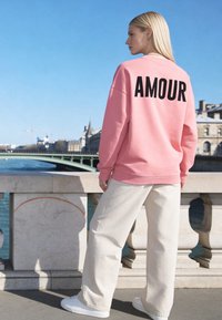 Femme aux longs cheveux blonds se tenant sur un pont en pierre, portant un sweat-shirt rose avec le texte "AMOUR", un pantalon blanc et des baskets au bord de la rivière.